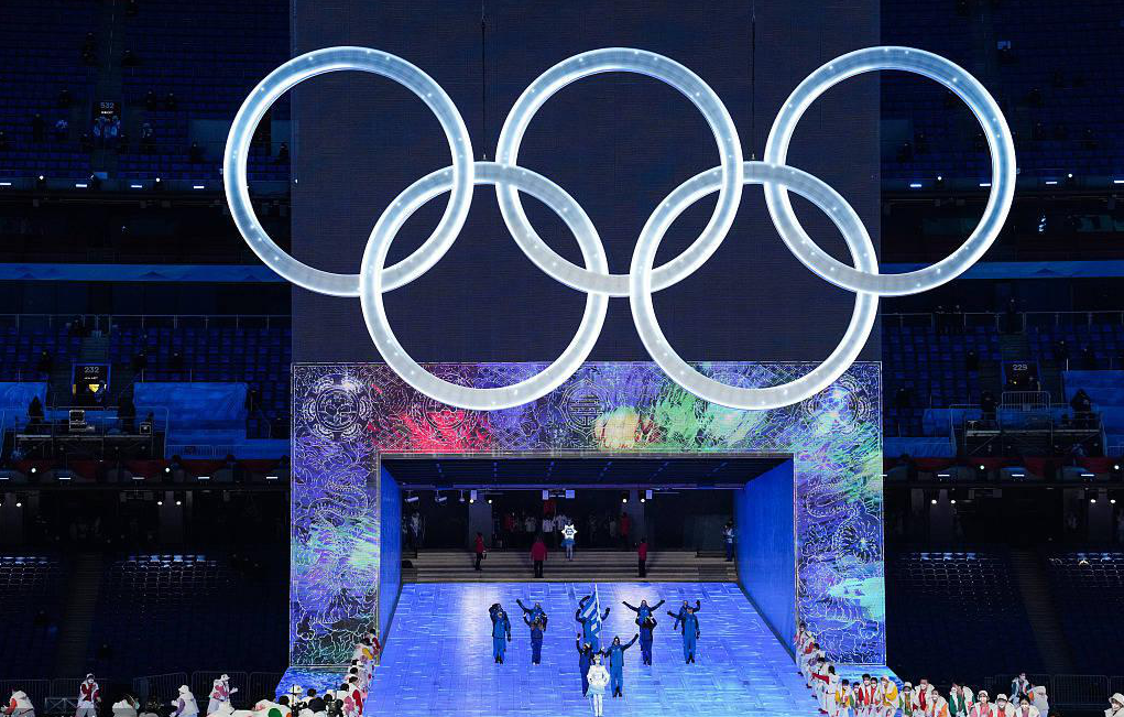 A group of athletes are greeting the audience, with the Olympic five-ring logo above them